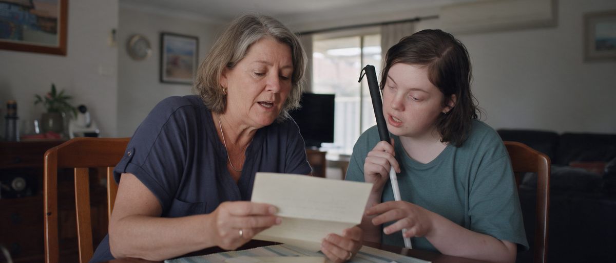 A mother reading a letter aloud to her adult daughter, who holds a white cane, at the dining table