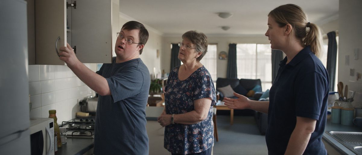 A man opening a kitchen cupboard as he and his mother are shown around a shared home by a support worker