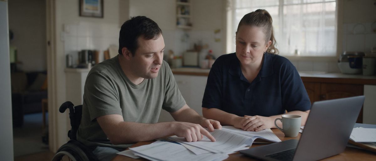 A man in a wheelchair and a support worker in a navy polo sit at a kitchen table, going through paperwork together with a laptop and a mug of tea