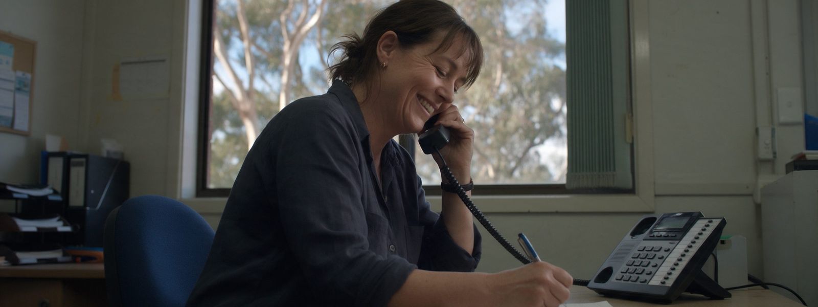 A woman smiling as she takes a phone call and writes a note at an office desk