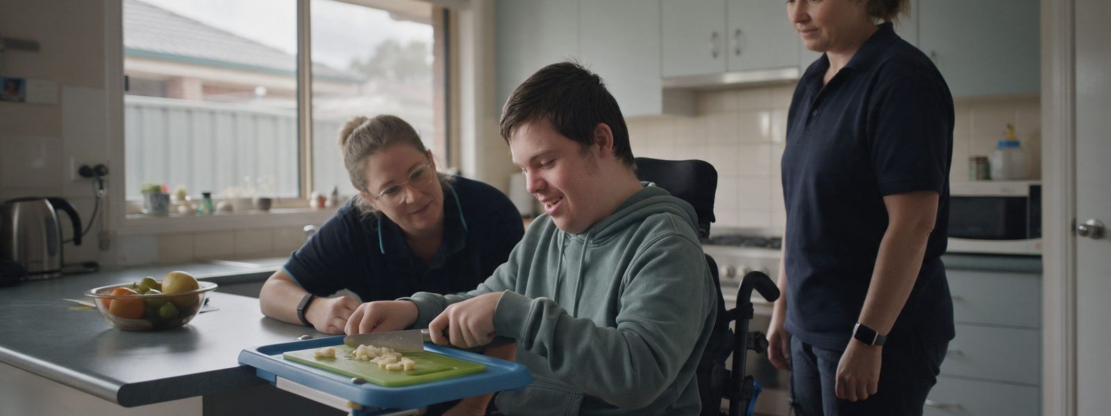 A young man in a wheelchair chopping food on an adapted board at his kitchen bench, with a therapist and a support worker beside him